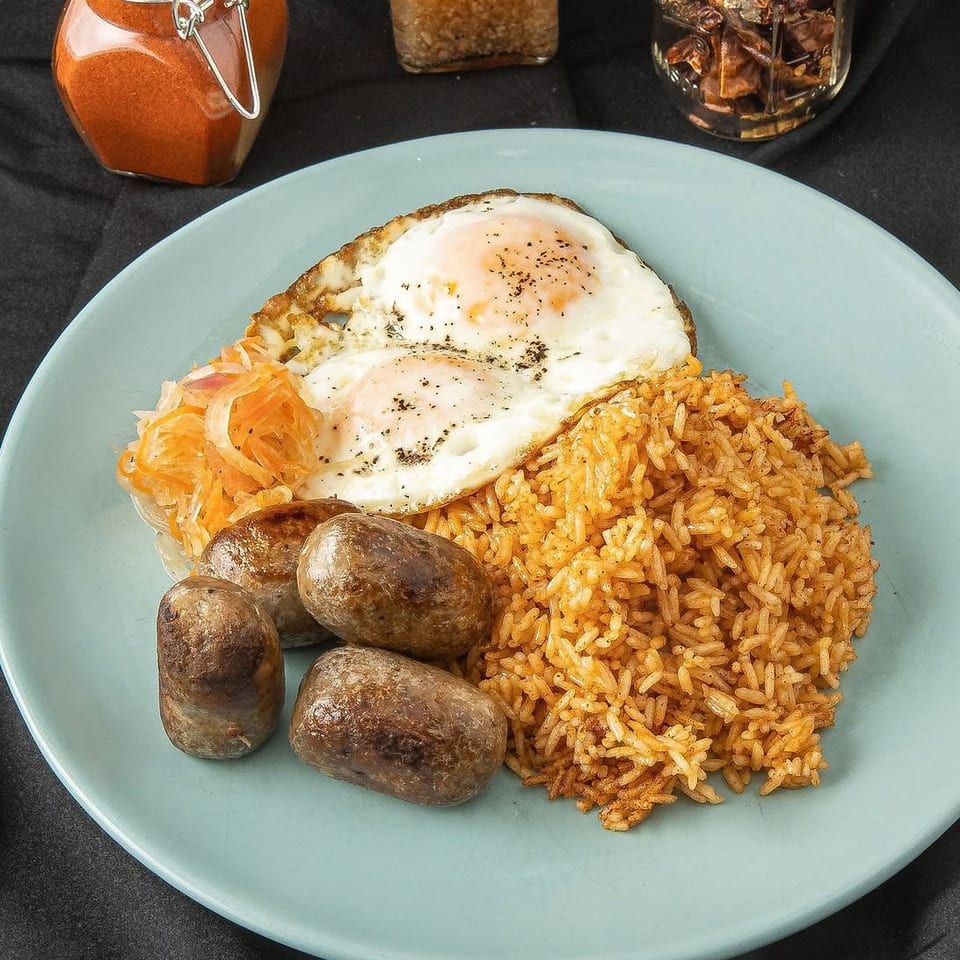 Plate of seasoned rice, fried eggs, roasted sausages, and shredded pickled vegetables with spice jars in the background.