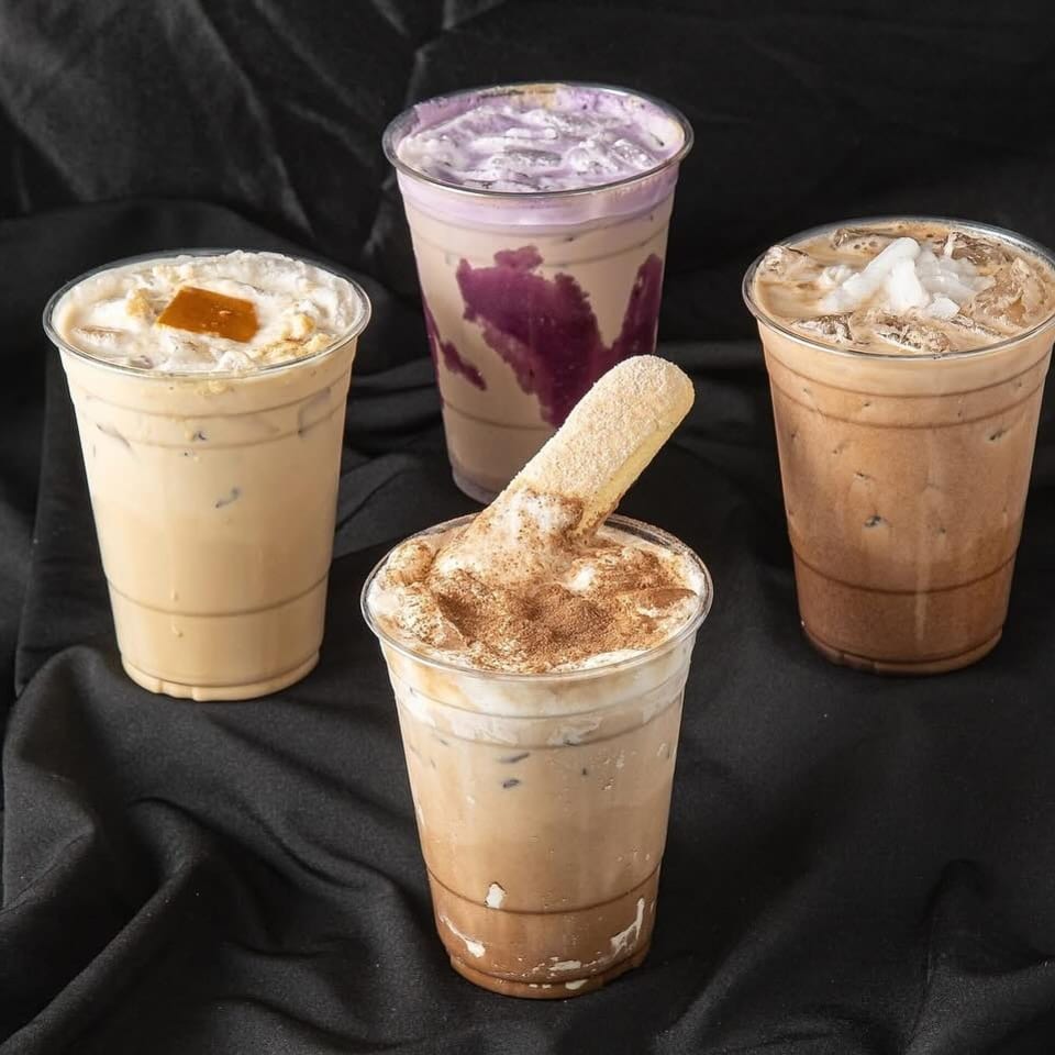 Four iced specialty drinks topped with whipped cream, spices, fruit, and a cinnamon-coated wafer on black fabric.
